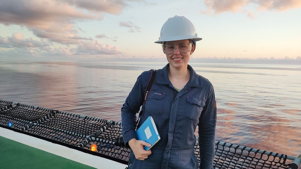 Photo of Monica on the helipad with a view of the ocean behind her at sunset. She is wearing protective gear and holding a notebook.